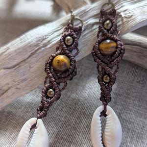 May include: Handmade macrame earrings with brown cord, tiger's eye beads, and small gold-coloured beads. Each earring has a white cowrie shell at the bottom. The earrings are displayed on a neutral grey surface.