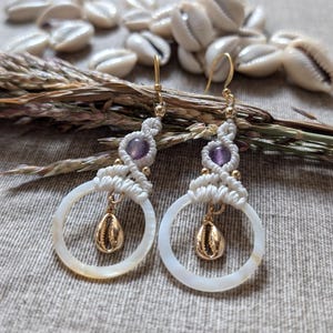 Può includere: Orecchini fatti a mano con macramè bianco, perline di ametista e dettagli color oro. Gli orecchini hanno un design circolare con un ciondolo a conchiglia dorata pendente. Lo sfondo include conchiglie e piante essiccate.