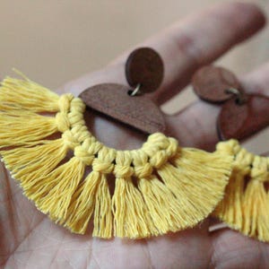 Può includere: Un paio di orecchini con nappe gialle con accenti in legno. Gli orecchini presentano un design a semicerchio con frange gialle annodate e piccoli borchie rotonde in legno marrone. Gli orecchini sono mostrati su uno sfondo neutro.