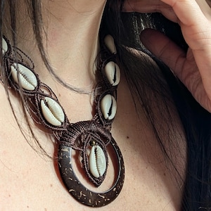 Könnte beinhalten: Eine braune Makramee-Halskette mit weißen Muscheln und einem dunkelbraunen Holzanhänger. Die Halskette wird um den Hals getragen und zeigt die komplizierten Knoten und natürlichen Materialien. Das Design ist böhmisch und strandnah.