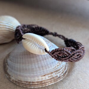 Könnte beinhalten: Ein braunes Makramee-Armband mit einer cremefarbenen Kaurimuschel. Das Armband wird auf einer Muschel präsentiert, mit einer weiteren Muschel im Hintergrund. Ein handgefertigtes Accessoire zum Thema Strand.