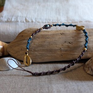 Peut inclure: Un bracelet macramé fait à la main avec un design de cordon bleu et marron. Le bracelet comporte des breloques coquillages dorées et de petites perles dorées. Le bracelet est présenté sur un morceau de bois flotté.