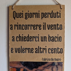 Könnte beinhalten: Rechteckiges Holzschild mit dem Text "Numero 1" oben. Das Schild zeigt italienischen Text und ein Gemälde eines Frauengesichts unten. Zum Aufhängen wird eine Juteschnur verwendet.