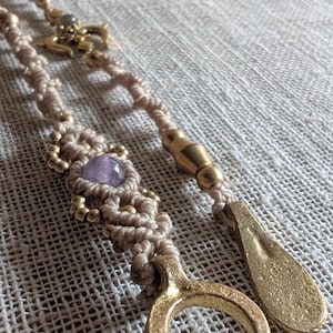 Op de afbeelding: Een macramé ketting met een lichtbeige koord, een centrale paarse edelsteen en kleine goudkleurige kralen. Het sieraad bevat een goudkleurige halve maan bedel en een druppelvormige hanger, afgewerkt met een metalen sluiting.