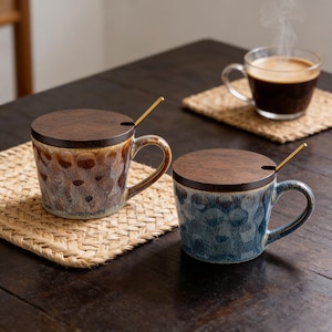 Op de afbeelding: Twee keramische mokken met houten deksels en gouden lepels staan op een donkere houten tafel. Eén mok is bruin en de andere blauw. Een helder glas koffie staat op de achtergrond op een geweven onderzetter.