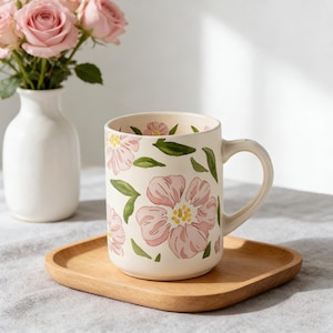 May include: Cream-colored ceramic mug with pink floral design and green leaves. The mug sits on a wooden tray. A white vase with pink roses is in the background.