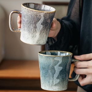 May include: Two ceramic mugs with a distinctive, textured design. One mug is held up, showing a gradient of grey and white, while the other is teal. Both mugs have a square handle and a rustic, artisanal aesthetic.