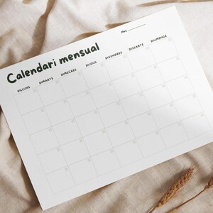 Op de afbeelding: Een witte maandkalender met de titel "Calendari mensual" in zwarte letters. De dagen van de week staan bovenaan vermeld. De kalender is leeg, met een raster van vierkanten voor elke dag. Twee aren tarwe staan in de rechter benedenhoek.