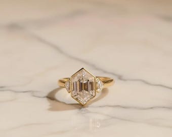 May include: A gold ring featuring a large, central, hexagon-cut clear gemstone set in a bezel, flanked by two smaller, triangular-cut clear side stones. The band is crafted from polished yellow gold, creating a sleek and refined appearance.