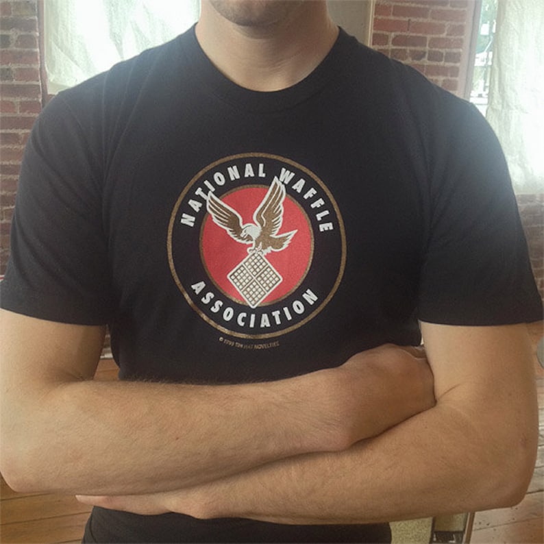 May include: A black t-shirt with a gold circle logo that says "National Waffle Association" with a red background and a white eagle holding a waffle. The logo also includes the text "&copy; 1999 TIN HAT NOVELTIES".