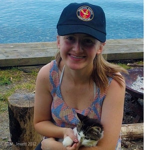 May include: A woman wearing a black baseball cap with the National Wildlife Federation logo, a colorful tank top, and holding a black and white cat.