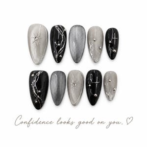 Puede incluir: Un juego de diez uñas postizas en tonos negro, plata y gris con puntas en forma de almendra. Los diseños presentan líneas metálicas plateadas, pequeños tachones plateados y detalles de estrellas. El texto "Confidence looks good on you." aparece impreso debajo.