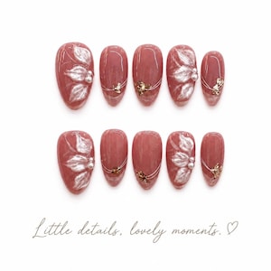 Puede incluir: Un juego de diez uñas postizas en tono rosa empolvado, con diseños florales blancos en relieve, pequeñas perlas blancas y finos detalles de alambre metálico dorado. Debajo aparece el texto "Little details, lovely moments." con un corazón.