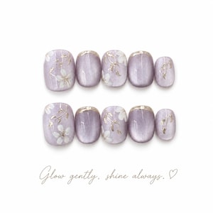 Puede incluir: Un juego de diez uñas postizas en un tono lavanda brillante, con detalles de flores blancas, hojas metálicas doradas y puntas con borde dorado. El texto "Glow gently, shine always." junto a un pequeño corazón aparece debajo de las uñas.