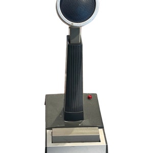 May include: A black and silver Shure microphone with a round, black speaker grille. The microphone is mounted on a gray base with a red button.