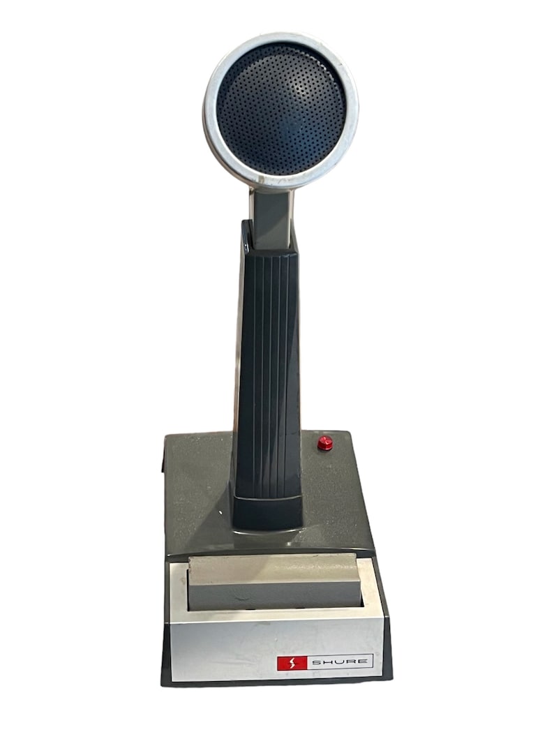 May include: A black and silver Shure microphone with a round, black speaker grille. The microphone is mounted on a gray base with a red button.