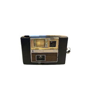 May include: A vintage Caravelle 747 camera. The camera has a black body with a silver flash and lens. The name "CARAVELLE" is printed in a silver font, and the number "747" is printed in a white font.