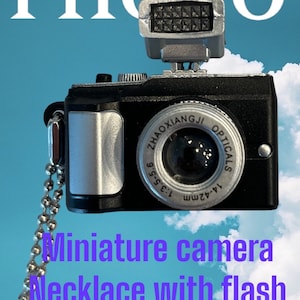 May include: A miniature black camera with a silver lens and a silver flash on top. The camera is attached to a silver chain, making it a necklace. The camera has the text "ZHAOXIANGJI OPTICALS 1:4.2mm 1:3.5-5.6" printed on the side.