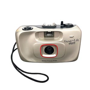 May include: A silver 35mm film camera with a black strap. The camera has a built-in flash and a red ring around the lens. The text "Friends for Life." and "LIFESCAN" is printed on the camera.
