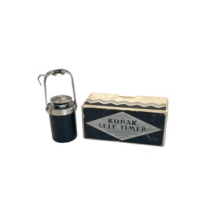 May include: A black Kodak self timer in its original box. The box is white with black and gray zig-zag patterns. The box has the text "Kodak Self Timer" printed on it.