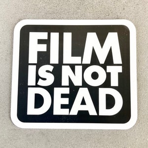 May include: Black and white sticker with the text "FILM IS NOT DEAD" in bold white letters on a black background.