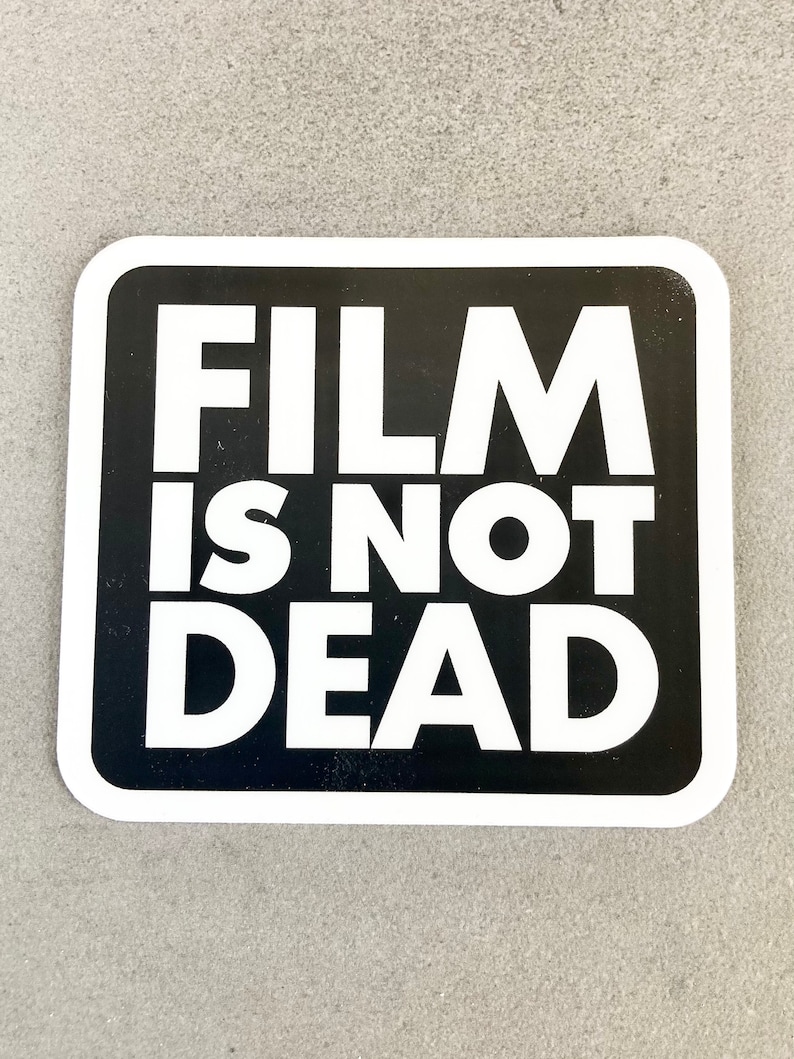 May include: Black and white sticker with the text "FILM IS NOT DEAD" in bold white letters on a black background.