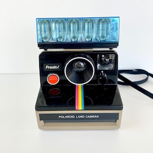 May include: A vintage Polaroid Land Camera, black with a tan base, features a rainbow stripe and the word "Presto!". It has a blue flash attachment and a black strap. The camera is a classic design.