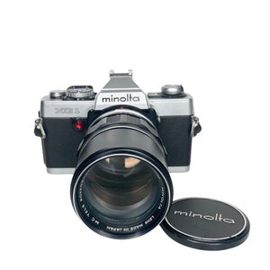 May include: A silver Minolta XG-1 35mm film camera with a black Minolta MC Tele Rokkor lens attached. The lens is labeled "Lens Made in Japan" and "MC Tele Rokkor". A black Minolta lens cap is also included.