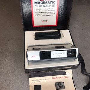 May include: A vintage silver and black Magimatic 210 pocket camera set in its original box. The camera has a red devil logo and the box has instructions for use.