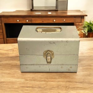 May include: A silver metal box with a brass handle and latch. The box has a slightly distressed look and is sitting on a wooden surface.