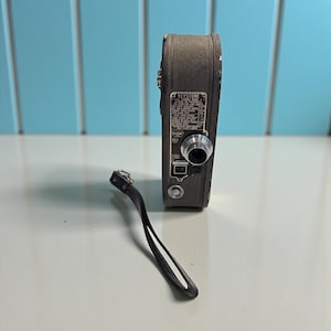May include: Vintage Keystone movie camera in a dark gray finish with a silver lens and a black wrist strap. The camera has a label with text. The camera is on a white surface with a blue background.