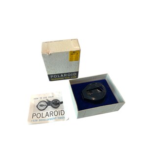 May include: A vintage Polaroid #128 Development Timer in its original box. The timer is black and has a white dial. The box is white with blue lettering and a yellow sticker.