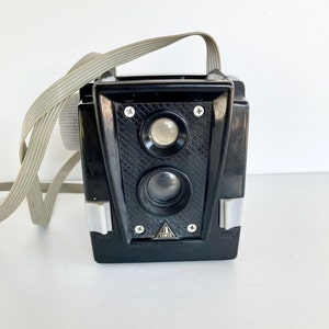 May include: A vintage black camera with a gray strap. The camera has two lenses and a textured front panel. Silver accents and screws are visible. The word "TOWER" is printed on the front. The camera is on a white surface.