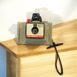 May include: A gray Polaroid Land Camera, model Big Swinger 360, with a black strap. The camera has a red and white label with the model name and a black lens.