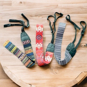 May include: Four camera straps with different colorful patterns. The straps are made of fabric and have adjustable lengths. The straps are on a wooden surface.