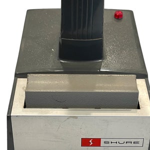 May include: A gray and silver microphone stand with a red button on the side. The stand has a black base with the word "SHURE" printed on it.