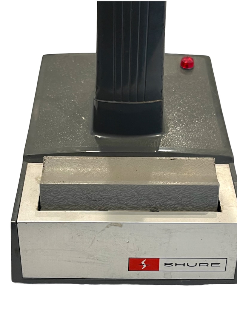 May include: A gray and silver microphone stand with a red button on the side. The stand has a black base with the word "SHURE" printed on it.