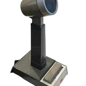 May include: A gray desktop microphone with a black base and a silver top. The microphone has a black grill and a silver ring around the top. The base has a silver plate with the word "Shure" printed on it.