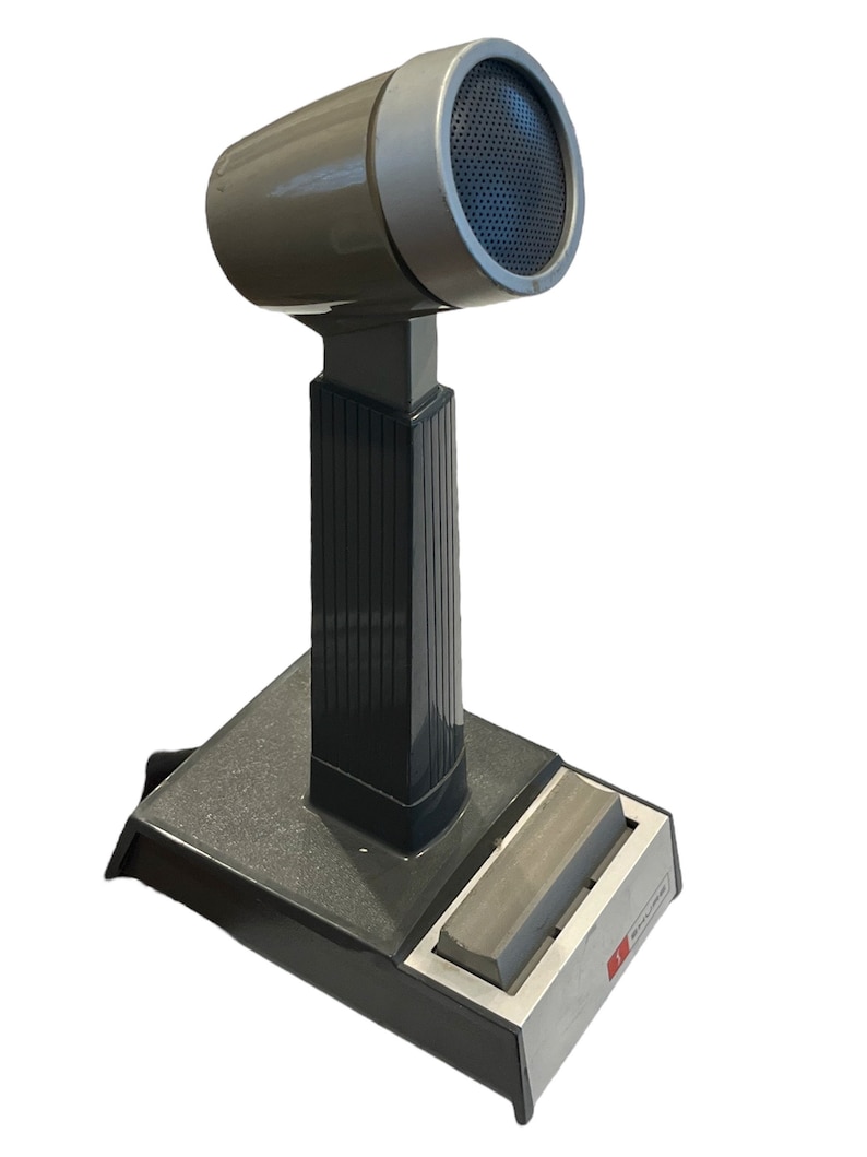 May include: A gray desktop microphone with a black base and a silver top. The microphone has a black grill and a silver ring around the top. The base has a silver plate with the word "Shure" printed on it.