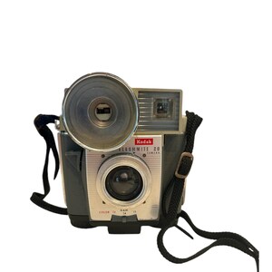 May include: A silver Kodak Flashmite 20 camera with a black strap. The camera has a built-in flash and a lens with a black ring around it. The camera has a black plastic body and a silver metal front.