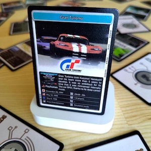 Może przedstawiać: Prostokątna karta kolekcjonerska z gry wideo Gran Turismo, przedstawiająca czerwony samochód sportowy, logo Gran Turismo oraz tekst opisujący rozwój gry, daty premiery i tryby rozgrywki, umieszczona na białej podstawce.