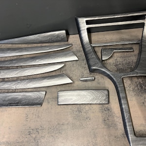 May include: A set of silver and black wood grain effect automotive interior trim components. The collection includes a centre console panel, dashboard trim, and smaller accent pieces. The items are displayed on a neutral surface.