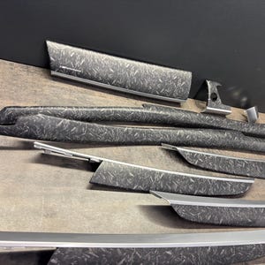 May include: A set of car interior trim components featuring a grey and black carbon fibre design. The pieces, of various shapes and sizes, have silver trim. They are displayed on a neutral surface.