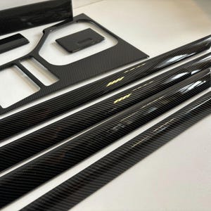 Puede incluir: Un conjunto de molduras interiores automotrices de fibra de carbono negra brillante. El juego incluye varias formas y tamaños, como tiras largas, paneles rectangulares y secciones contorneadas, todas con un patrón tejido.