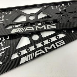 Może przedstawiać: Dwie czarne ramki tablic rejestracyjnych z włókna węglowego z literami "AMG" w kolorze srebrnym. Ramki mają wzór o fakturze splotu i różne wycięte kształty. Przeznaczone do użytku motoryzacyjnego.