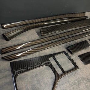 Peut inclure: Un ensemble de garnitures intérieures automobiles présentant un motif en fibre de carbone brillant. La collection comprend plusieurs bandes de tableau de bord allongées, un contour de levier de vitesse de console centrale et des panneaux rectangulaires.