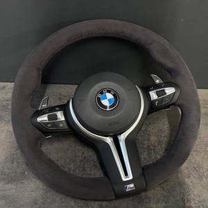Könnte beinhalten: Ein dunkelgraues BMW-Lenkrad aus Wildleder mit einem zentralen runden BMW-Logo, integrierten schwarzen Bedientasten an den horizontalen Speichen, silbernen Schaltwippen und einem kleinen M-Emblem unten an der mittleren Speiche.