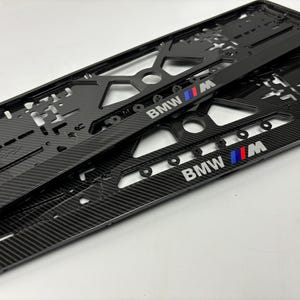 Może przedstawiać: Dwie czarne ramki tablic rejestracyjnych z włókna węglowego z logo BMW M w kolorze białym, niebieskim i czerwonym. Ramki mają teksturowany wzór i wycięte sekcje.