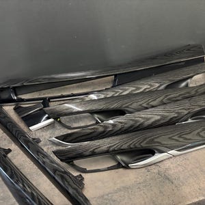 May include: A selection of car interior trim components featuring a wood-effect finish. The parts are black with a grey wood grain and silver detailing. They are displayed on a grey surface.