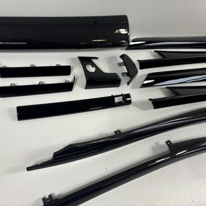 May include: An assortment of glossy black car components, including a panel with carbon fibre weave and the text "M Performance". Various trim pieces and components are arranged on a white surface, showcasing a sleek, modern aesthetic.